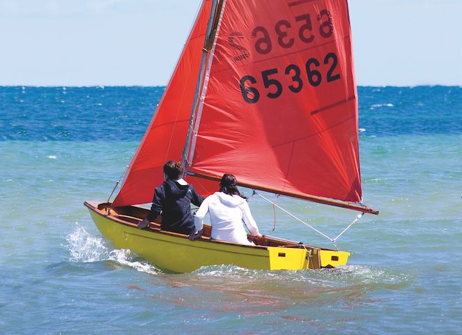 two men in a small sailboat on the water with red sails and numbers on it