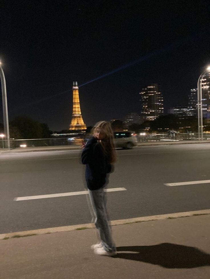 a woman is standing on the sidewalk in front of the eiffel tower at night