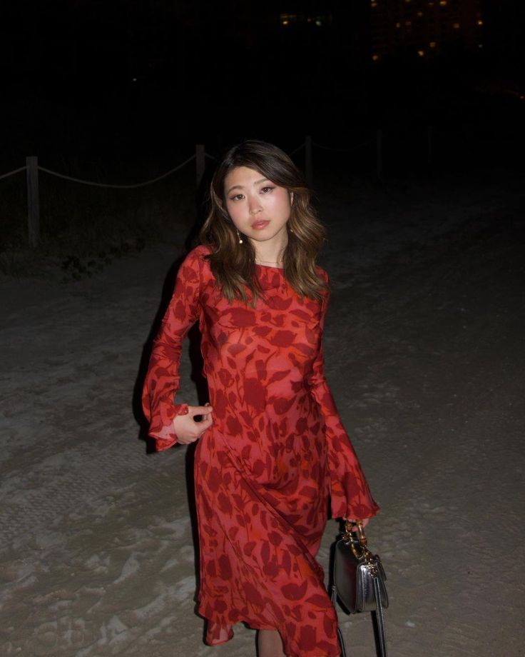 a woman in a red dress is standing on the beach at night with her purse