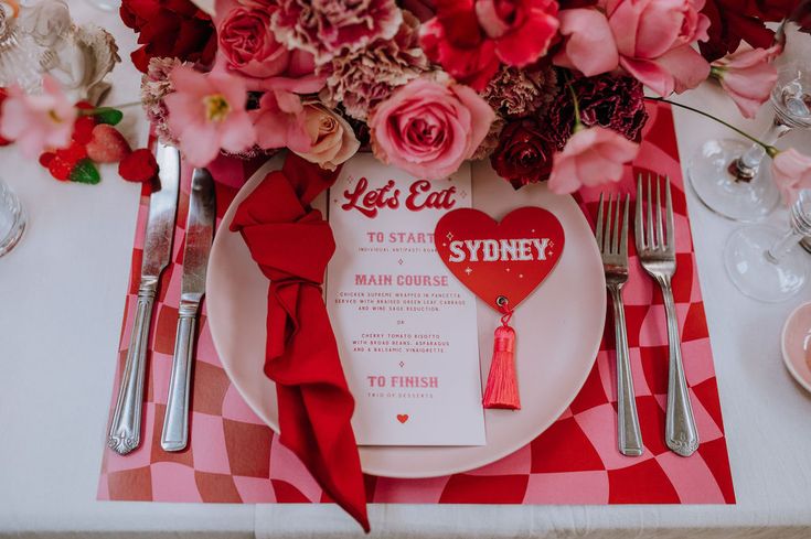 A table setting with red and pink checkerboard placemat, a pink plate with a red napkin, menu and red heart shaped name card with a red tassel with red and pink flowers at the top of the image Wedding Poker Table, Pink And Red Retro Wedding, Pink Red Table Setting, Valentine Wedding Theme, Valentines Themed Wedding, Pink And Red Table Setting, Disco Valentines Day, Valentines Elopement, Valentines Wedding Theme