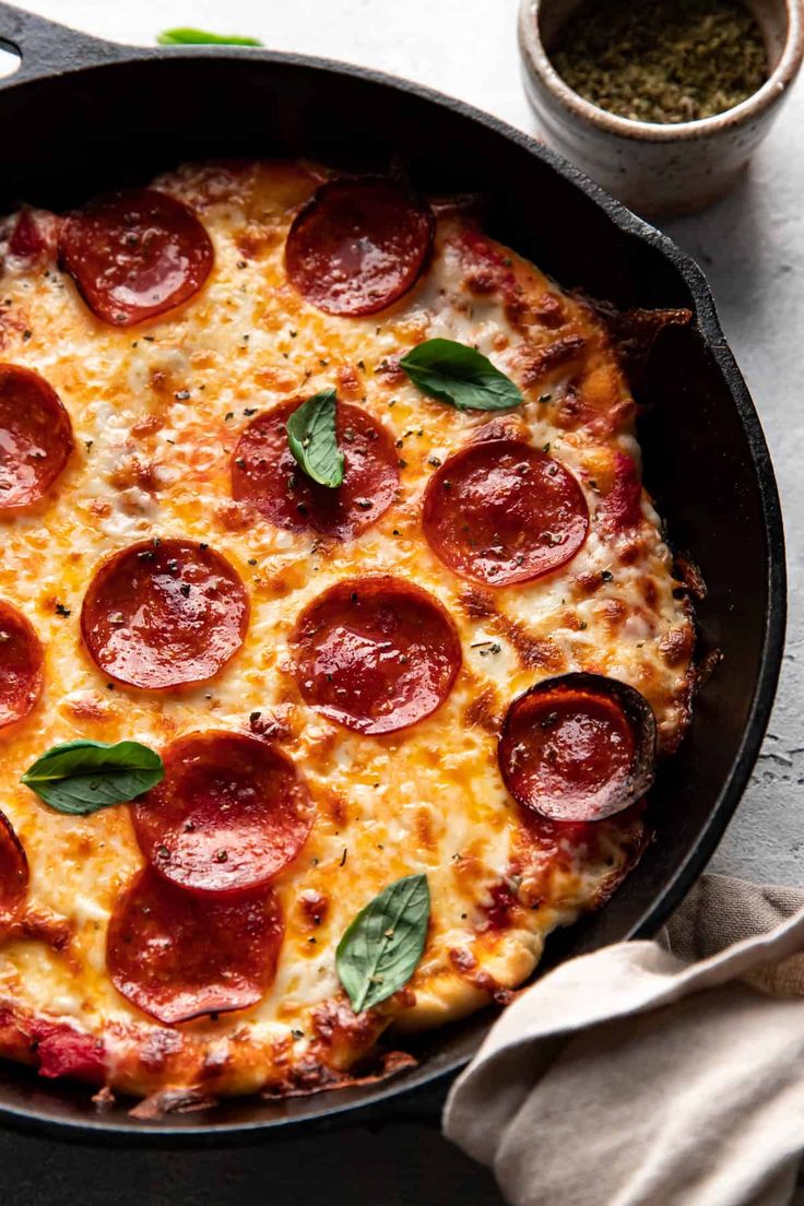 a pepperoni pizza in a cast iron skillet with basil leaves on the side