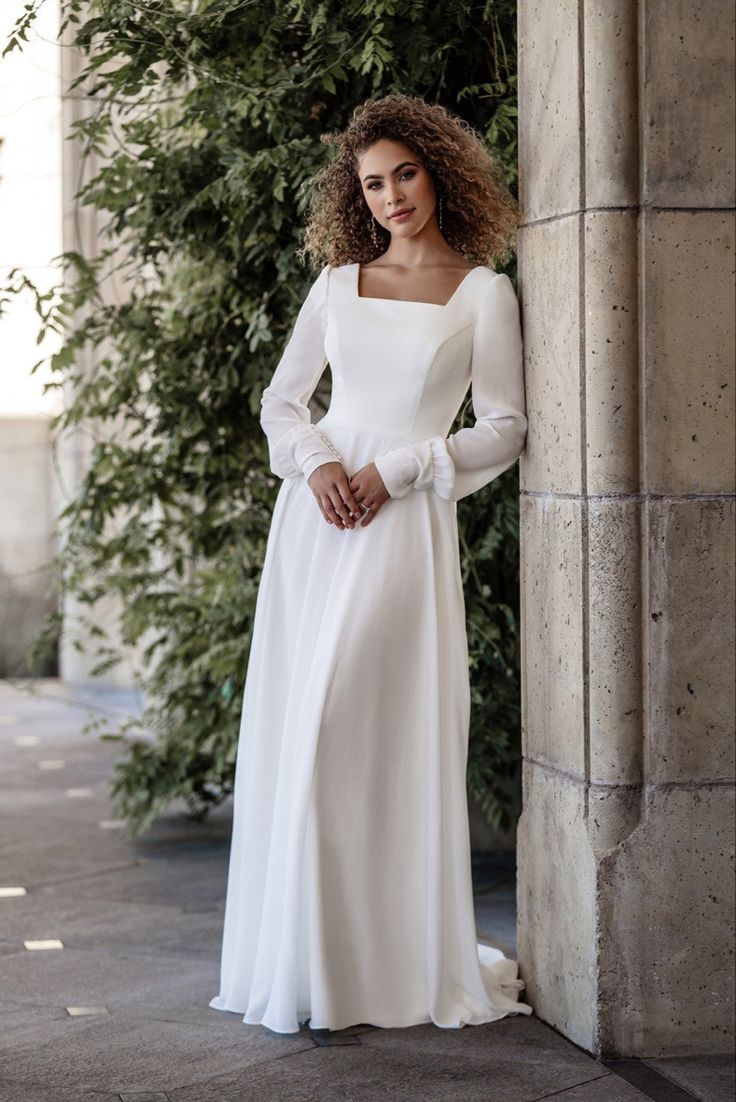 a woman in a white dress leaning against a wall with her hands on her hips