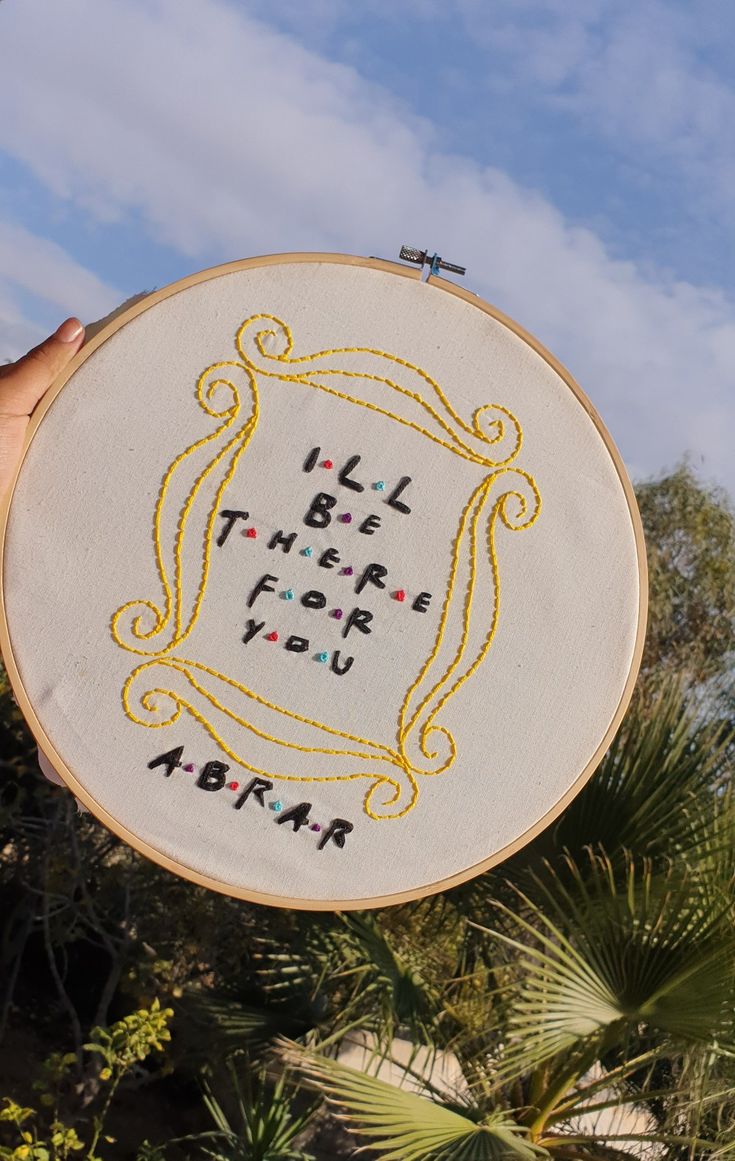 a person holding up a white embroidery hoop with words written in black and yellow on it