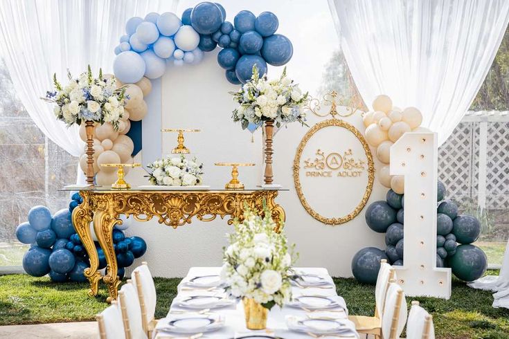 a table set up with blue and white balloons, gold chairs, and a cake