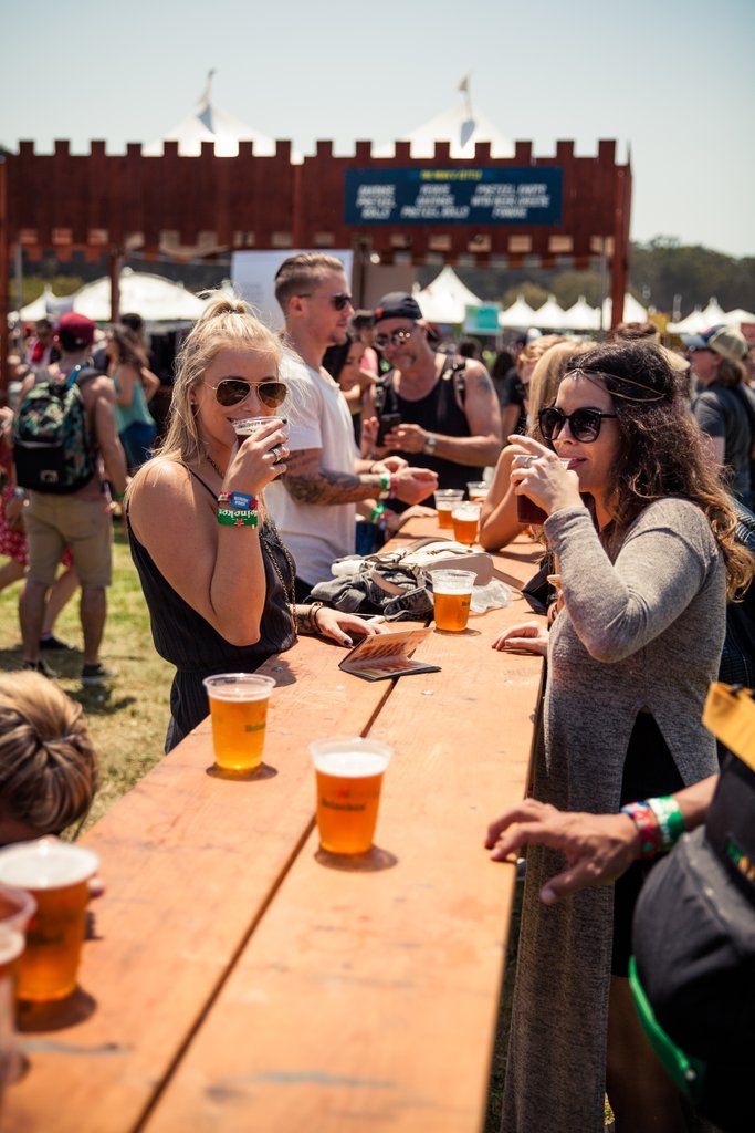 a group of people sitting around a wooden table with beers in front of them at an outdoor event