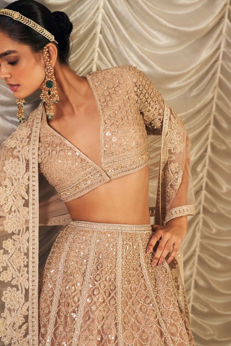 Exude opulence in the champagne net embroidered blouse, paired with a matching net embroidered lehenga and dupatta. The blouse features intricate embroidery in a soft champagne hue, offering a subtle yet luxurious sparkle. The lehenga, crafted from the same elegant net fabric, is richly embellished to complement the blouse. The net embroidered dupatta completes the look with its delicate, ethereal charm, making this ensemble perfect for any grand occasion. Beige Zari Work Pre-draped Saree For Wedding, Beige Pre-draped Saree With Zari Work For Wedding, Beige Sets With Mirror Work For Reception, Festive Anarkali Beige Choli, Bollywood Style Beige Lehenga For Party, Wedding Party Wear Blouse With Intricate Embroidery, Anarkali Style Beige Choli For Reception, Elegant Semi-stitched Cream Lehenga, Elegant Beige Choli With Traditional Drape