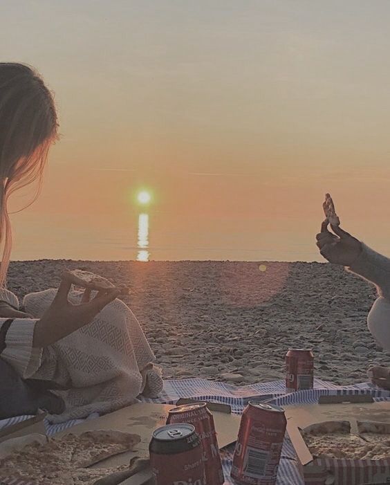 two people sitting at a picnic table eating pizza and drinking beer on the beach with the sun setting in the background