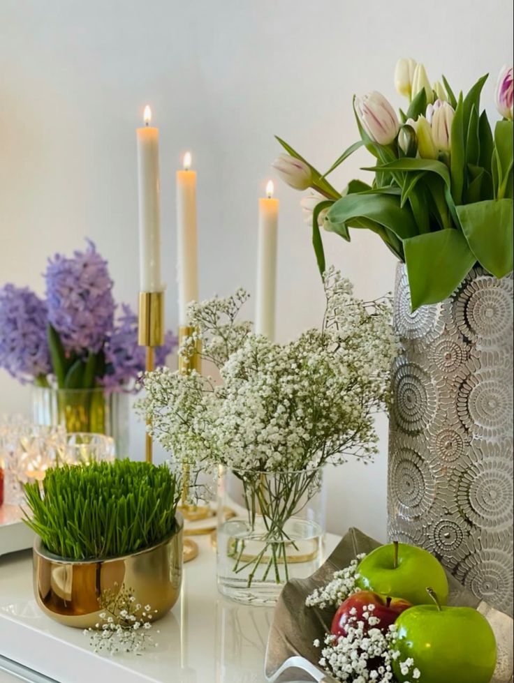 flowers and candles are sitting on a table with vases filled with flowers, fruit and greenery