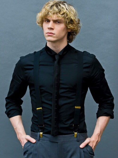 a young man with blonde hair wearing a black dress shirt and suspenders, standing in front of a gray wall