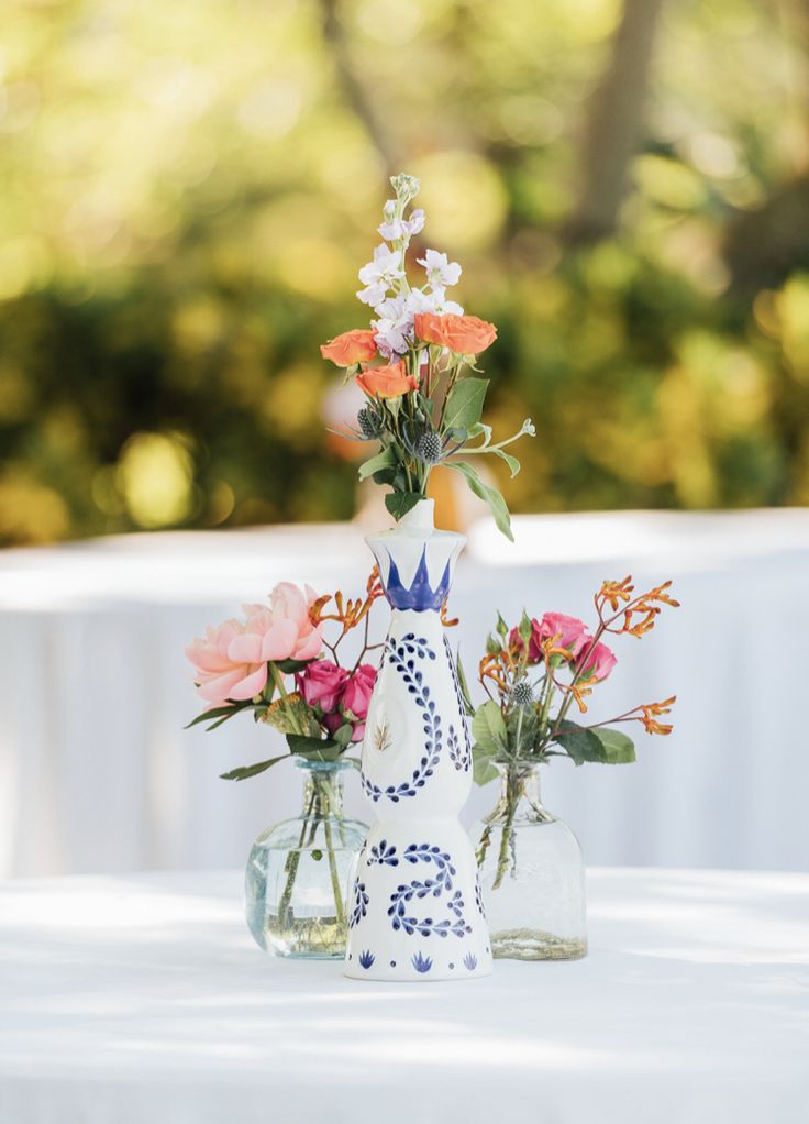 two vases with flowers are sitting on a white table cloth covered tablecloth in front of some trees