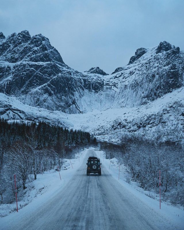 a truck driving down a snow covered road in the middle of mountains with trees on both sides