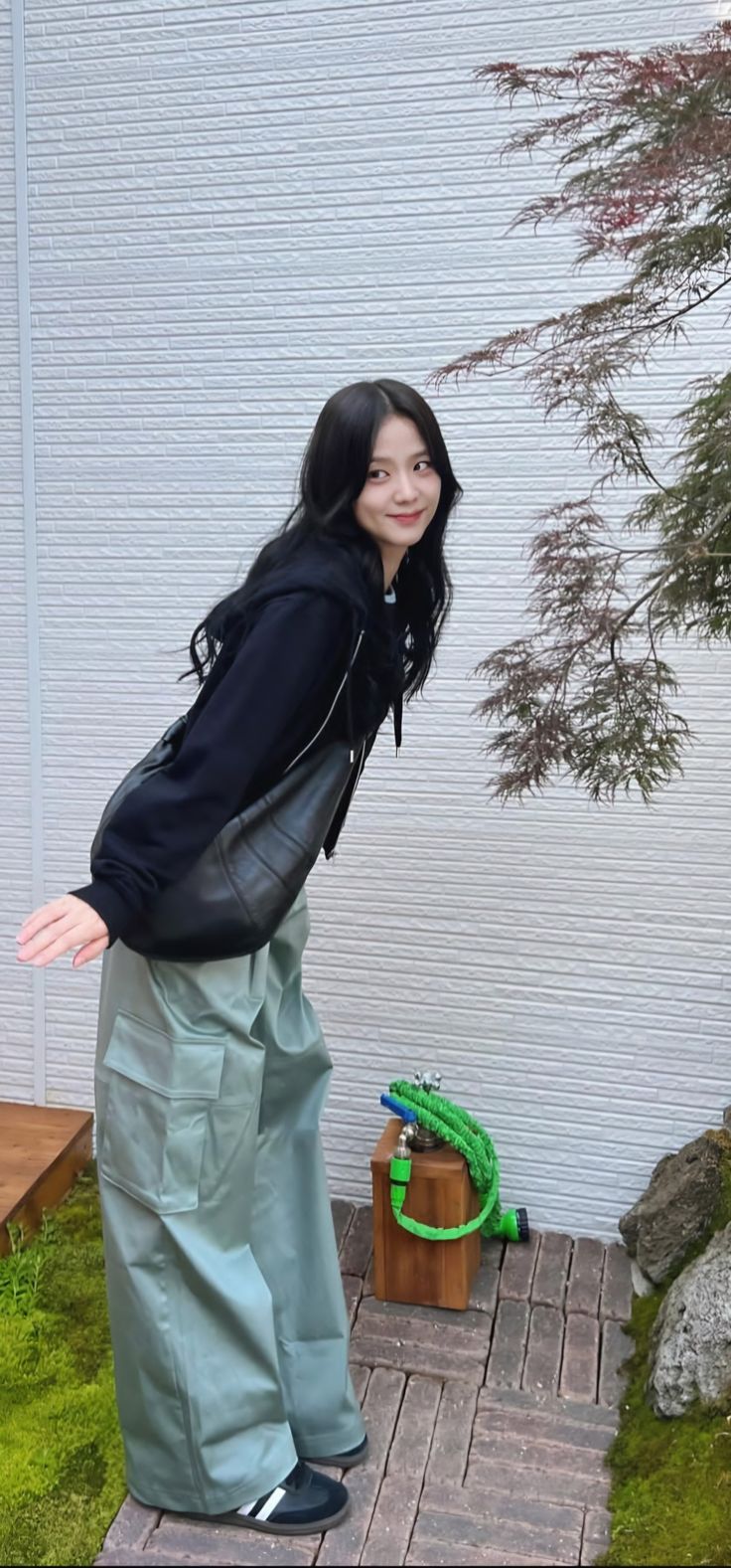 a woman standing in front of a white wall next to a green plant and tree