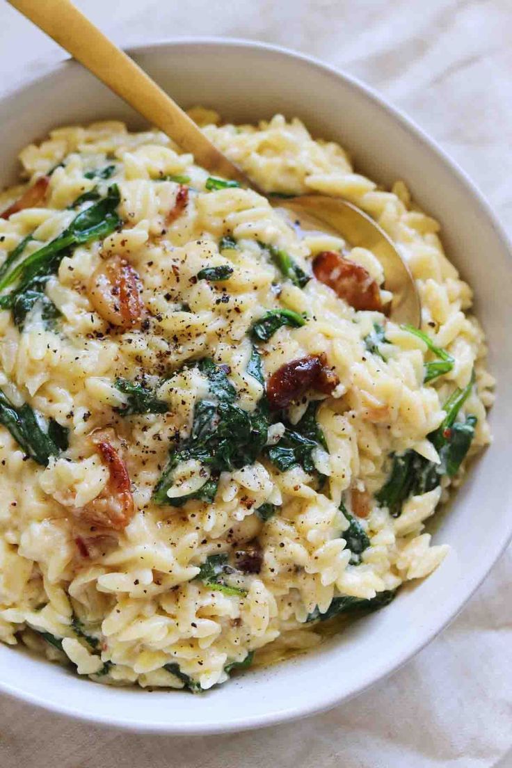 a bowl filled with pasta and spinach on top of a white table cloth next to a wooden spoon