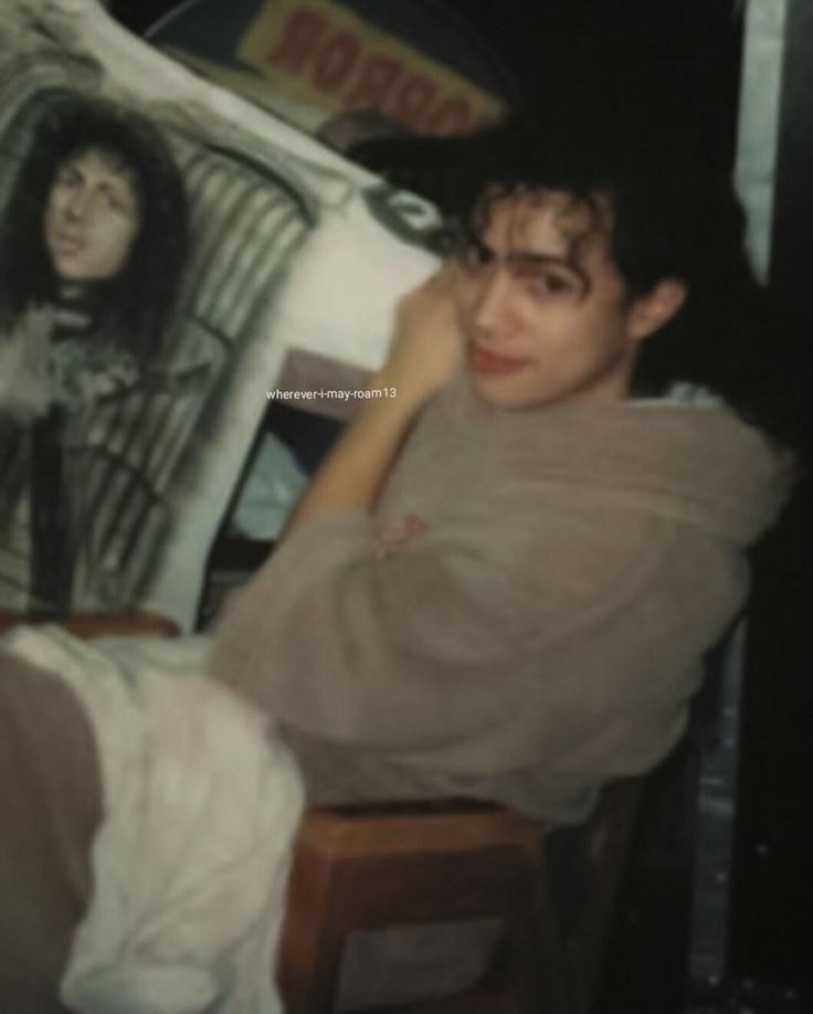 a young man sitting on top of a wooden chair in front of a painting with an image of a woman