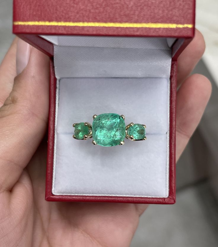 A Colombian emerald three stone ring. This bespoke ring design is crafted in gleaming 14K yellow gold, this ring features a 6.55-carat cushion cut in its center. Oval emeralds are flanked on the side creating an array of green. Simply gorgeous and one of a kind! Setting: Three-Stone Metal Purity: 14K Yellow Gold Center Stone: Emerald Shape: Cushion Cut Estimated Center Weight: 6.55-Carats Clarity: Semi-Transparent Color: Green Luster: Very Good Origin: Colombia Treatments: Natural Side Stones: E Heirloom Style Three Stone Green Emerald Ring, Heirloom Green Emerald Ring With Three Stones, Heirloom Green Three Stone Ring, Green Emerald Three Stone Ring In 14k Gold, Green Three-stone Emerald Ring For Anniversary, Luxury Three Stone Emerald Ring Gift, Green Emerald Three Stone Ring For Anniversary, Green Three Stone Emerald Ring For Anniversary, Luxury Oval Emerald Ring With Three Stones