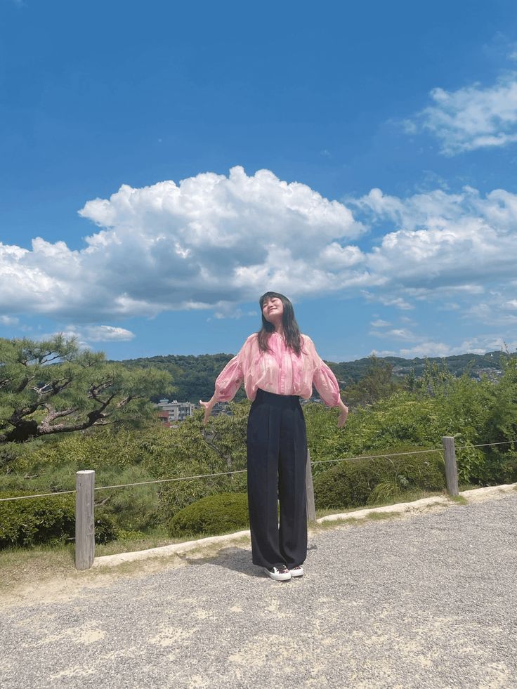 a woman standing on top of a dirt road next to a lush green forest under a blue sky with white clouds