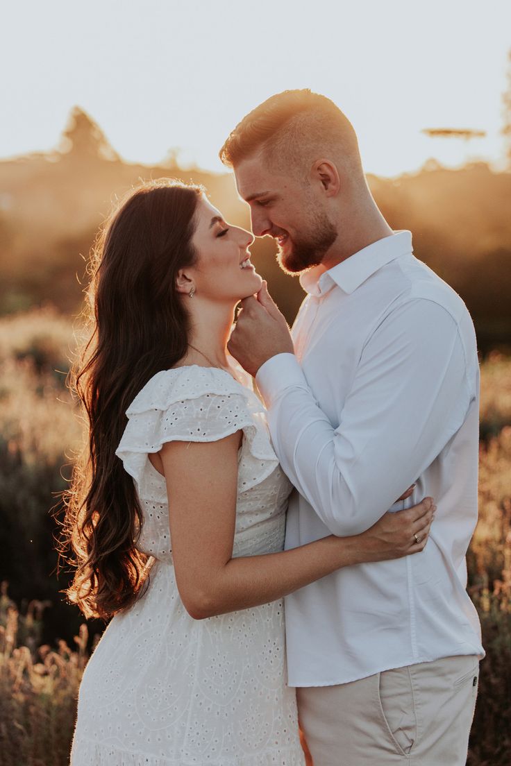 a man and woman standing next to each other in front of tall grass at sunset