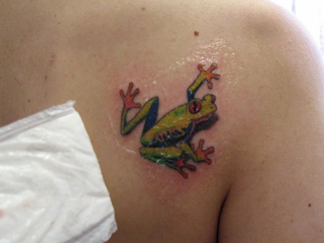 a green frog tattoo on the back of a woman's left shoulder and chest