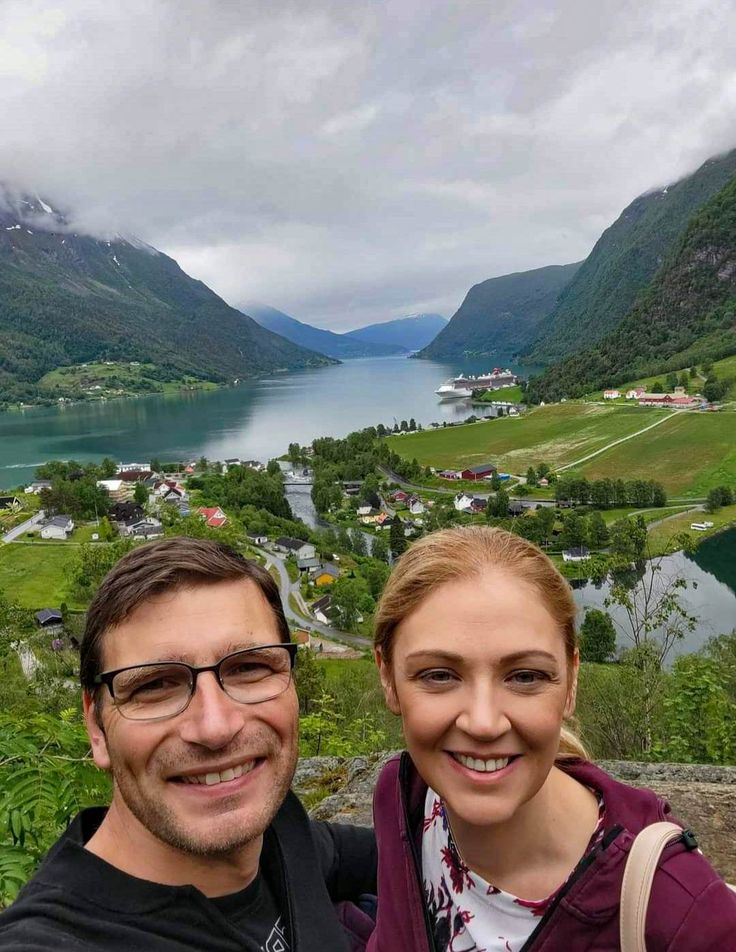 a man and woman standing next to each other on top of a hill near a lake