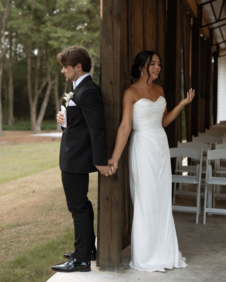 a bride and groom standing next to each other in front of a wooden structure holding hands