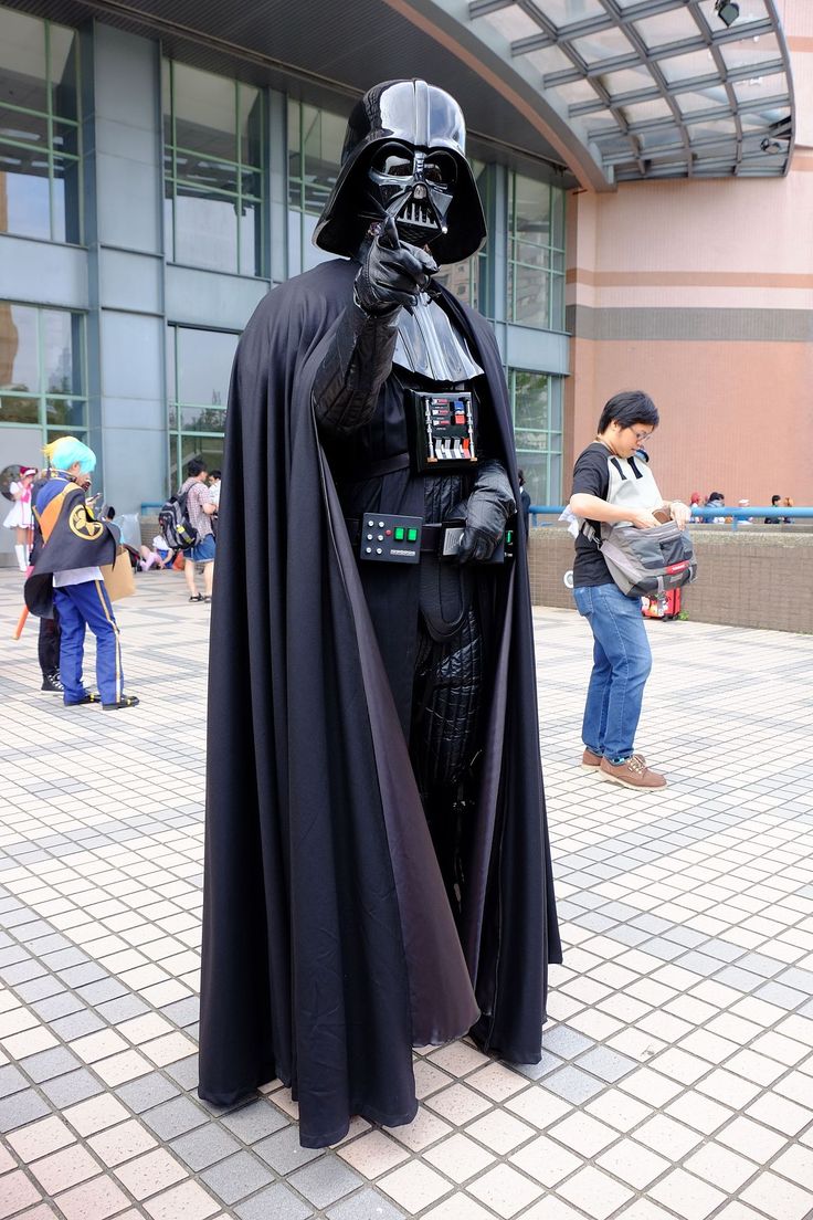 a man dressed as darth vader stands in front of a building with people standing around