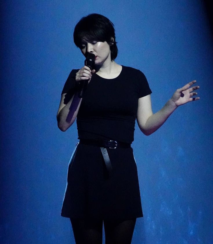 a woman standing in front of a blue wall holding a microphone to her mouth while wearing black