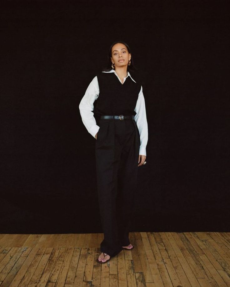 a woman standing in front of a black background wearing a white shirt and black pants