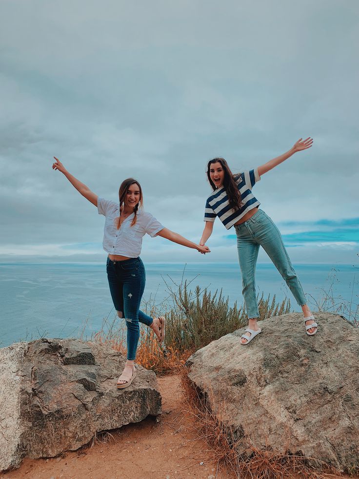 two young women standing on top of rocks near the ocean with their arms in the air