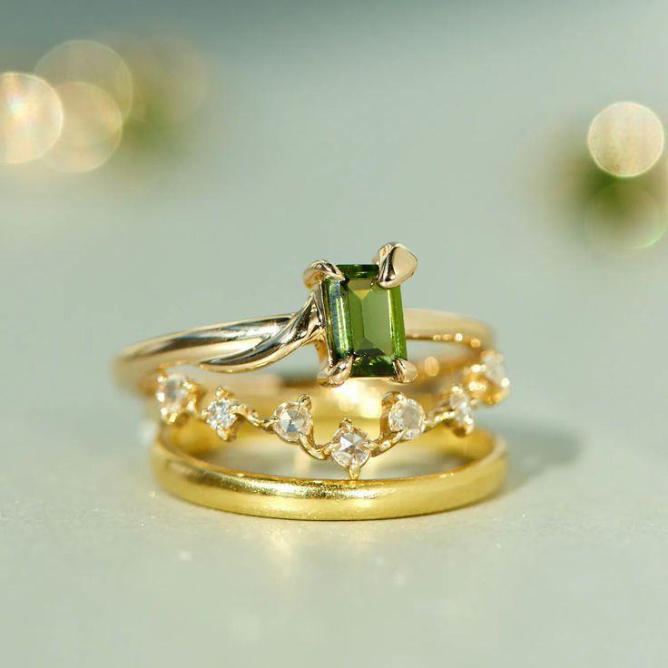 A stack of rings on a table. From top to bottom they're: a tourmaline ring with a central green stone with a gold band that slithers around it. The second is a curved band with dainty diamonds. The bottom is a solid gold rounded band. In the background are glowing out of focus stones. Fine Ring, Pearl And Diamond Earrings, Living Things, Green A, Tourmaline Ring, Tennis Bracelet Diamond, Engagement Ring Wedding Band, Green Tourmaline, Diamond Bracelets