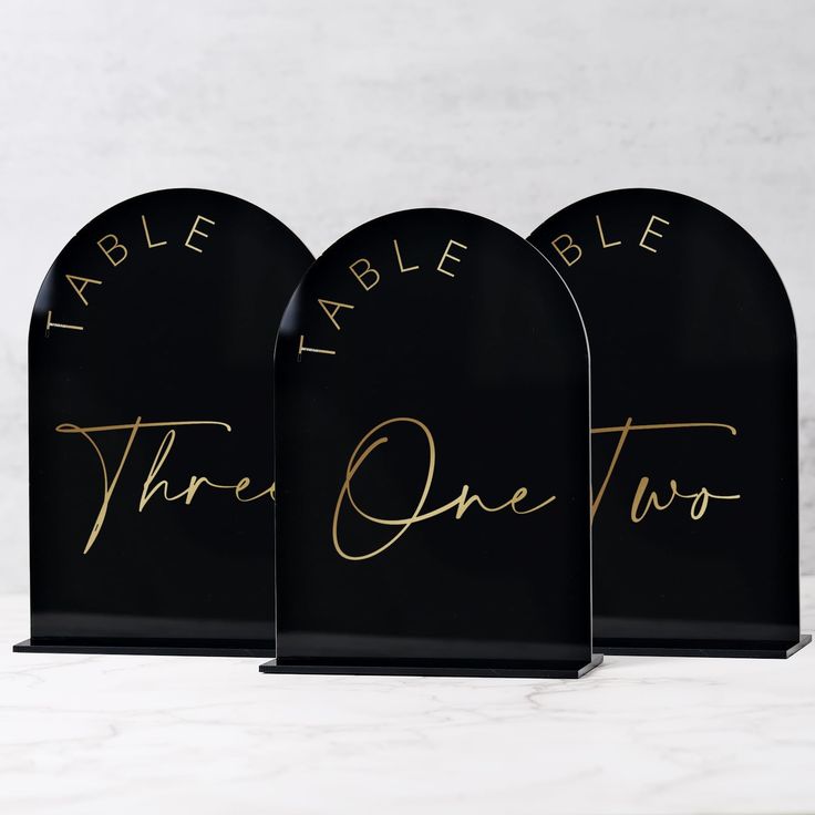 three black acrylic signs with gold writing on them that say table, table, and the one two