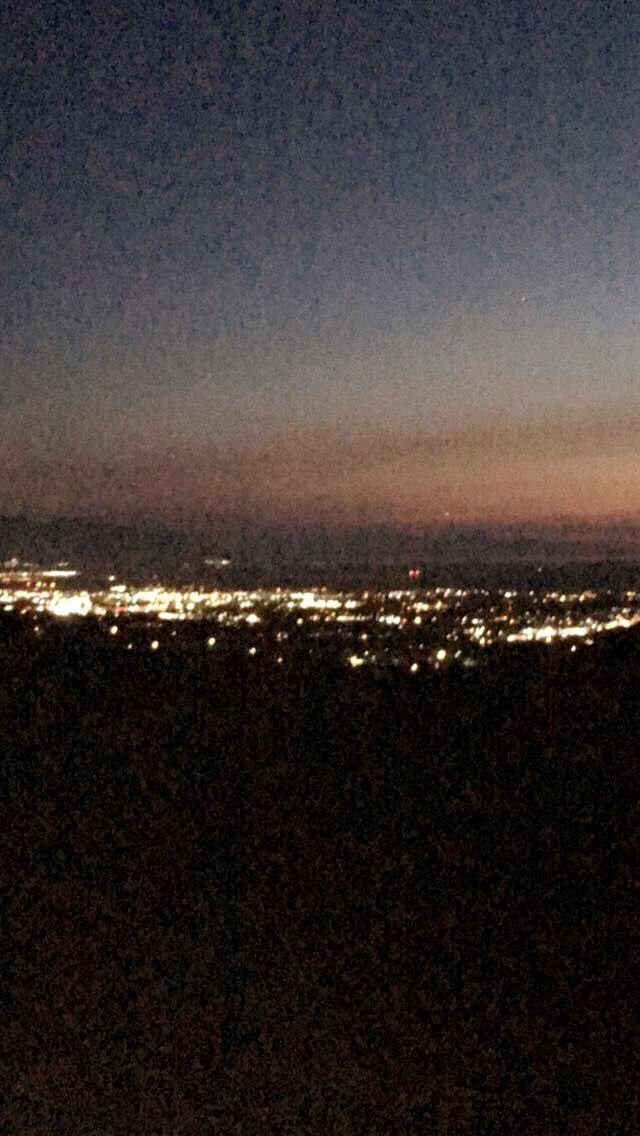 the city lights are lit up in the night sky from atop a hill at dusk