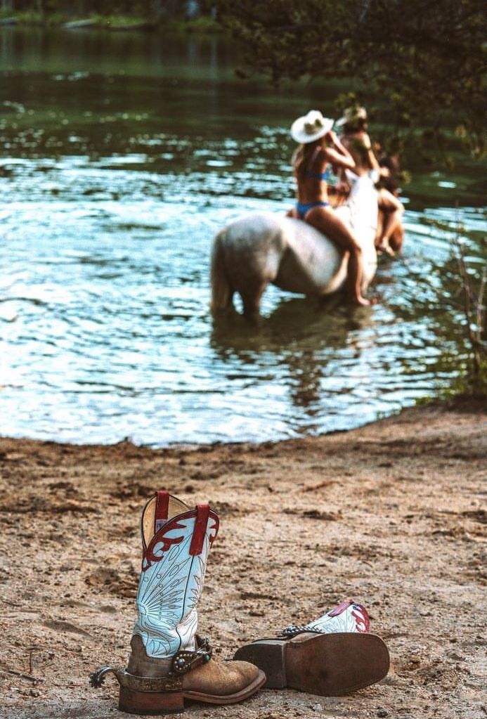 two people riding horses in the water with cowboy boots on the ground next to them