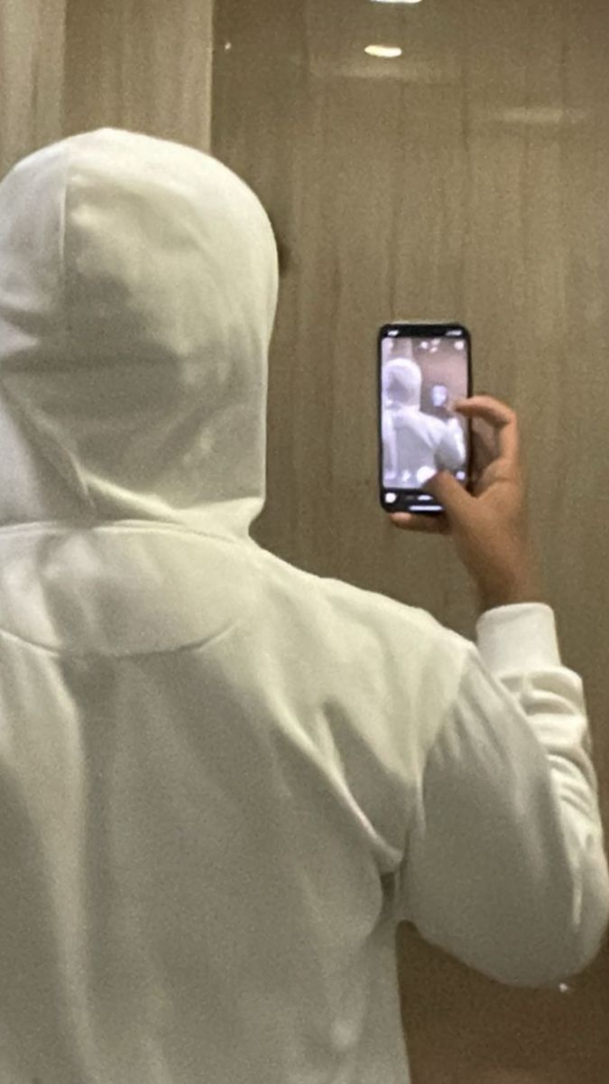 a person in a white hoodie taking a selfie with a cell phone while standing in front of a mirror