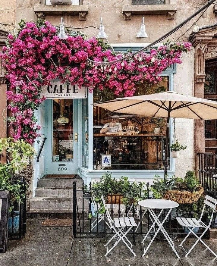 an outdoor cafe with pink flowers growing over the tables and chairs on the sidewalk in front of it