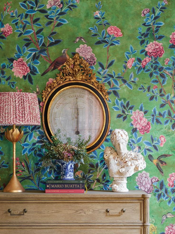 a dresser with a mirror, lamp and vase on it in front of a floral wallpaper