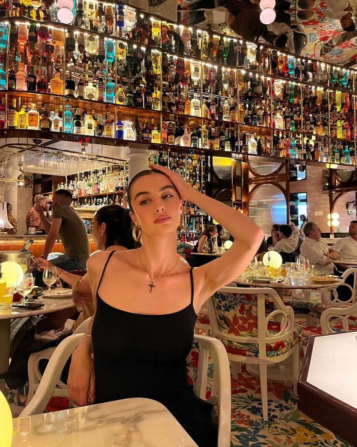 a woman sitting at a table in front of a bar with many bottles on the wall