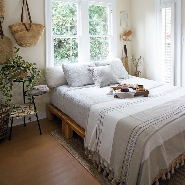 a white bed sitting under a window next to a wooden table with two baskets on top of it