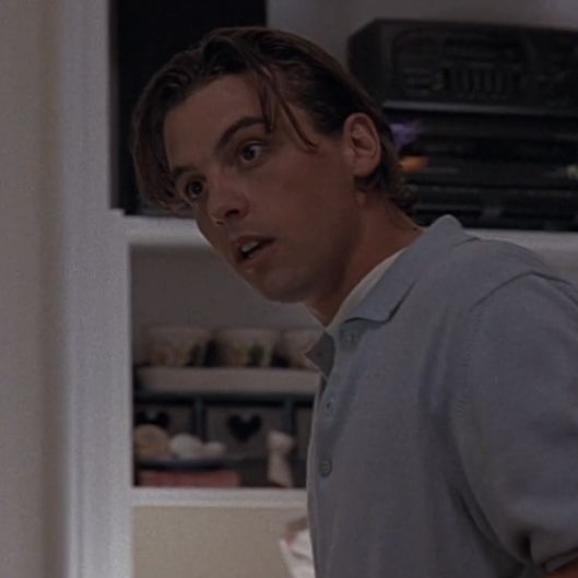 a man in grey shirt standing next to a shelf with dishes on it and an open microwave