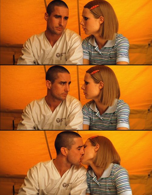 a man and woman sitting next to each other in front of a yellow wall kissing