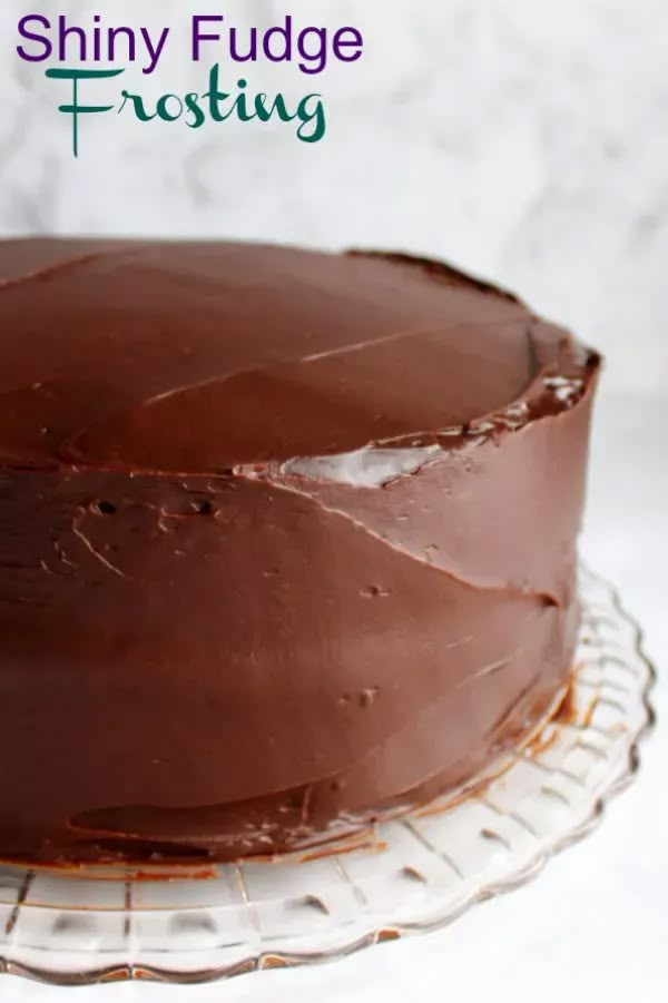 a chocolate frosted cake sitting on top of a glass plate with the words shiny fudge frosting