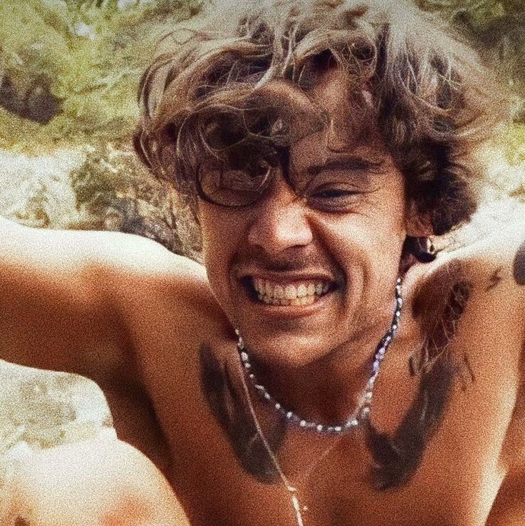 a shirtless man sitting on the beach with his hands behind his head and tattoos on his chest