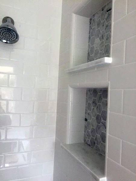 a white tiled bathroom with shelves on the wall and shower head mounted to the side