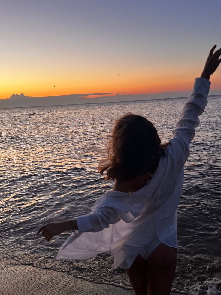 a woman standing on the beach with her arms in the air as the sun sets