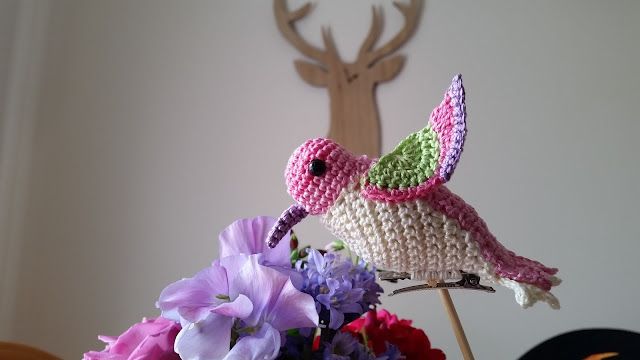 two crocheted hummingbirds sitting on top of a table next to purple flowers