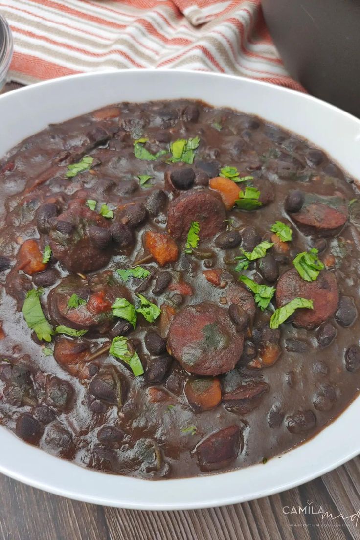 a white bowl filled with beans and meat on top of a wooden table next to a cup of coffee