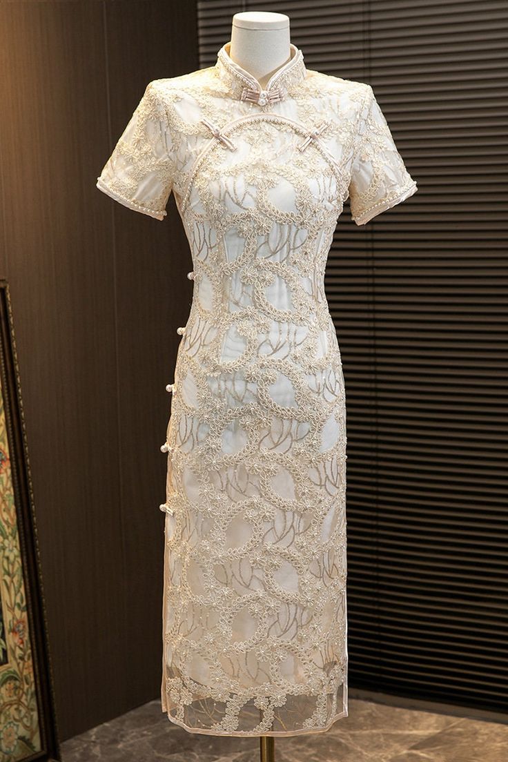 Introducing the stunning short sleeves white lace qipao with delicate floral appliques and faux pearls, a true masterpiece in traditional Chinese fashion. This gorgeous dress is crafted from high-quality lace material that feels soft and comfortable against the skin.The dress features intricate floral appliques that ar Elegant Lace Dress With Floral Embroidery And Short Sleeves, Lace Dress With Pearl Embroidery For Ceremony, Elegant White Ao Dai For Formal Occasions, Ceremony Lace Dresses With Pearl Embroidery, Elegant Short Sleeve Embroidered Lace Dress, Elegant White Dress With Pearl Buttons, Short Sleeve Lace Dress With Floral Embroidery, Elegant White Short Sleeve Lace Dress, White Lace Dress With Short Sleeves