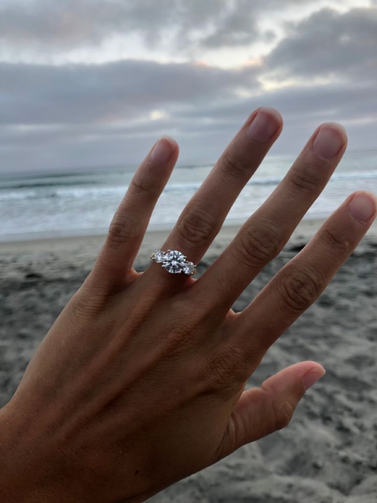 a person's hand with a diamond ring on the beach in front of an ocean