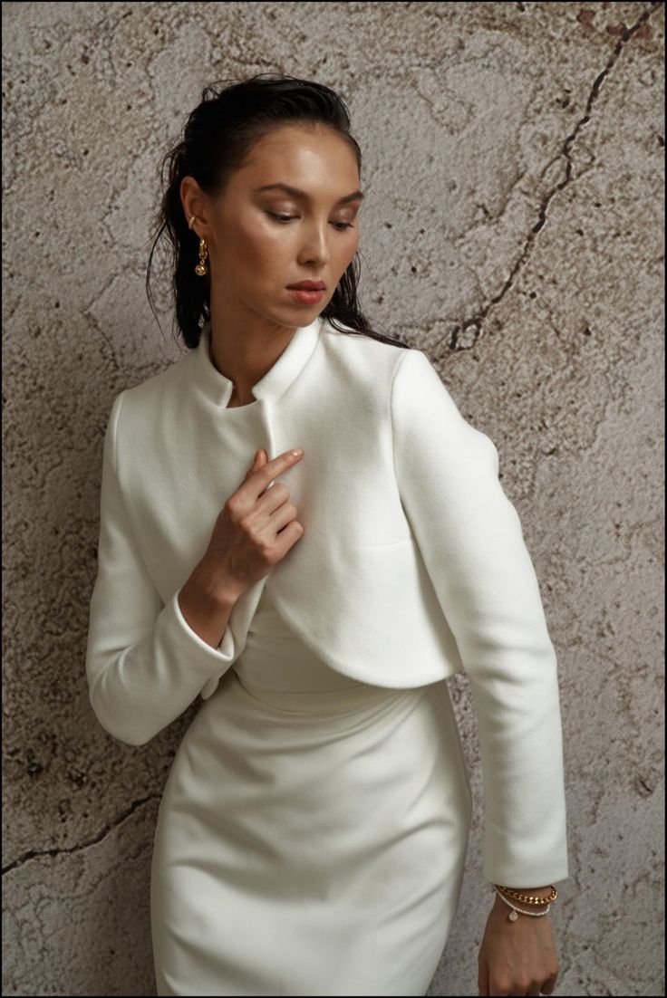 a woman wearing a white dress and jacket standing next to a wall with her hands on her chest