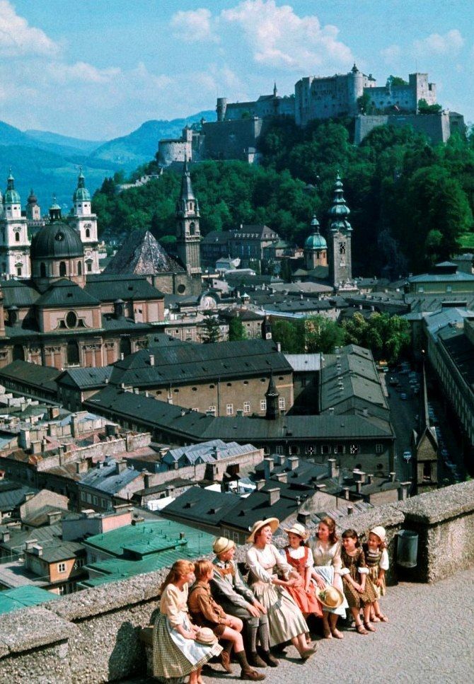 several people sitting on the edge of a wall overlooking a city with mountains in the background