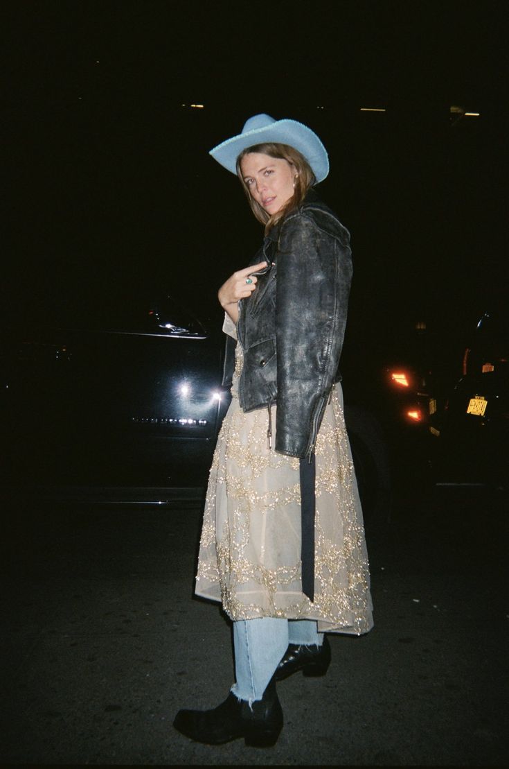 a woman wearing a hat and jacket standing in the dark with her hand on her hip